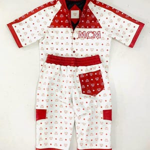 Pode incluir: Um conjunto de duas peças com uma camisa de mangas curtas e calções a condizer. O conjunto é branco com um padrão de diamante vermelho repetido e acabamentos vermelhos. A camisa tem a inscrição "MCM". O conjunto é apresentado sobre um fundo branco.