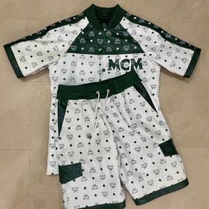 May include: White and green matching athletic set featuring a short-sleeved jacket and shorts. The jacket and shorts are patterned with a repeating logo design and trimmed with green accents. The jacket has the letters "MCM" on the front.