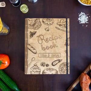 Op de afbeelding: Een houten receptenboek met een bruine kaft en een zwarte rug. De kaft is versierd met verschillende voedsel-illustraties, waaronder een brood, een kom pasta en een tros druiven. De tekst "Recipe Book" is gedrukt in een decoratief lettertype op de kaft, samen met de namen "Ella & John".