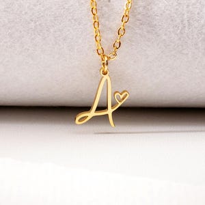 May include: A gold-toned necklace featuring a delicate chain and a pendant shaped like the cursive letter A, which includes a small, open heart detail attached to the right side of the letter. The metal has a polished, reflective finish.
