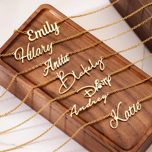 Peut inclure: Colliers dorés avec des noms, présentés sur un plateau en bois. Chaque collier affiche un nom différent en écriture cursive : Emily, Hilary, Anita, Blakely, Dany, Audrey et Katie. Les colliers sont présentés dans une boîte en bois.