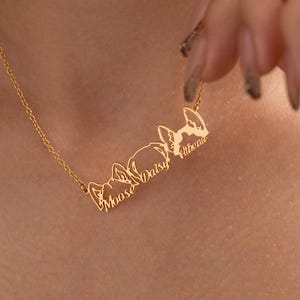 May include: A gold-colored necklace with a delicate chain. The pendant features three dog silhouettes with the names "Moose", "Daisy", and "Athena" engraved below each dog. The necklace is being held up by a person.