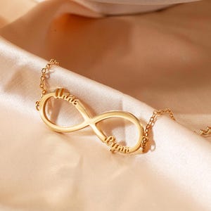May include: A gold-coloured infinity necklace with the names "Anna" and "Ryan" engraved on it. The necklace is displayed on a peach-coloured satin fabric, with a white ceramic bowl in the background.