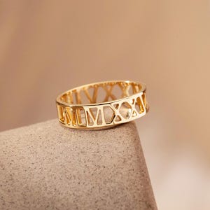 May include: A gold-colored ring with Roman numerals cut out around the band. The ring is resting on a gray, textured surface, with a blurred, neutral-toned background.