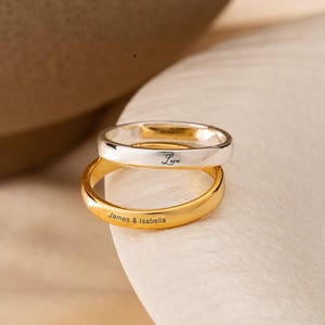 Peut inclure: Deux bagues superposées, une en argent et une en or. La bague en argent porte l'inscription "Love". La bague en or porte l'inscription "James & Isabella". Les bagues sont posées sur une surface blanche.