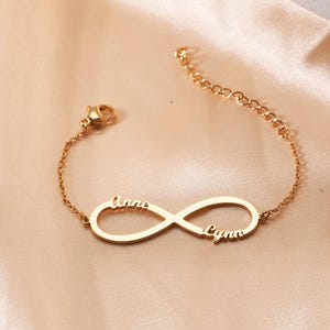 Könnte beinhalten: Goldfarbenes Armband mit einem Unendlichkeitssymbol und den eingravierten Namen "Anne" und "Lynn". Das Armband hat eine zarte Kette und einen Verschluss und liegt auf einer satinartigen Oberfläche. Ein personalisiertes Schmuckstück.