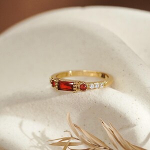 May include: A gold ring featuring a central baguette-cut red gemstone flanked by two smaller round red stones and a series of tiny clear stones on each side. The band has a polished finish and a delicate, thin profile with intricate metal detailing.