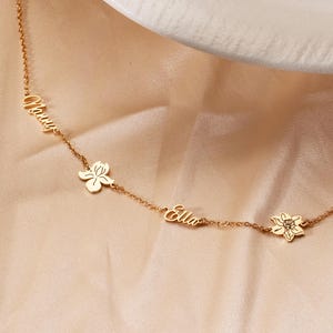 Puede incluir: Un delicado collar dorado con los nombres "Nancy" y "Ella" grabados. El collar presenta dos colgantes de flores y una pequeña gema. La cadena es fina y elegante, perfecta para el uso diario.