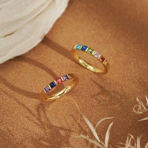May include: Two gold-toned rings, each featuring a row of small, square-cut multicoloured gemstones. The stones are set in a channel style along the top half of the band, displaying a vibrant spectrum of colours including blue, red, and purple.