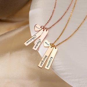 Op de afbeelding: Twee metalen kettingen met dunne schakels. Een roségouden ketting heeft twee rechthoekige hangers gegraveerd met de namen Andrew en Sophia, plus een klein hartje met de tekst Forever. Een gouden ketting heeft twee rechthoekige hangers met Morgan en Kristina, plus een klein hartje met de tekst Mom.