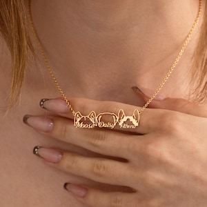 May include: A gold-colored necklace featuring three stylized dog head pendants, each with a name engraved: Moose, Daisy, and Athena. The necklace is displayed against a skin-toned background, highlighting the delicate chain and pendants.