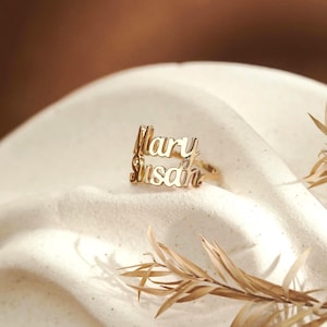 May include: A gold-toned ring featuring two stacked lines of cursive script text. The top line displays the name Mary and the bottom line displays the name Susan. The metal has a polished, reflective finish and the letters are connected to the band.