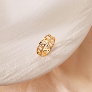 Op de afbeelding: Een goudkleurige ring met een uitgesneden ontwerp bestaande uit herhalende Romeinse cijfers rondom de gehele omtrek van de band. Het metaal heeft een gepolijste afwerking, wat zorgt voor een reflecterend oppervlak dat de open structuur benadrukt.