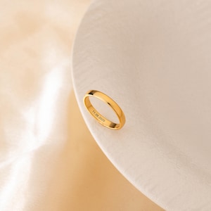 May include: A gold ring with the engraved date "11.08.2020" is shown on a white surface. The ring has a smooth, polished finish and a simple design. The background is a soft, neutral colour.