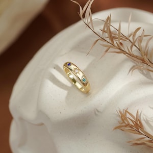 Könnte beinhalten: Ein goldener Bandring mit drei kleinen, bündig gefassten Edelsteinen in Blau, Transparent und Türkis. Jeder Stein ist von einem eingravierten Sternenmuster umgeben, was ein himmlisches Design auf der polierten, abgerundeten Metalloberfläche erzeugt.