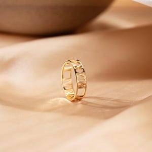 Puede incluir: Un anillo dorado con números romanos grabados alrededor de la banda. El anillo se muestra sobre una superficie de color melocotón. El diseño es abierto, permitiendo que la luz pase a través de los números.