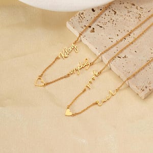May include: Gold-coloured personalised name necklaces. Each necklace features a delicate chain with a cursive name pendant. One necklace has a heart charm. The necklaces are displayed on a neutral-toned surface.