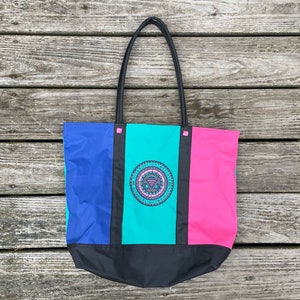 May include: A color-blocked tote bag featuring vertical panels of blue, teal, and pink, separated by black strips. The center teal panel displays a circular logo with the text GITANO SLIGHTWEIGHTS. It has black shoulder straps and a black base.