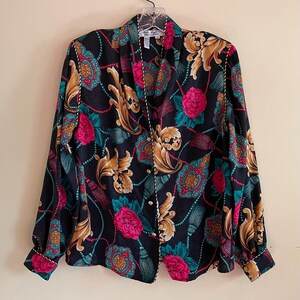 May include: A long-sleeved blouse with a dark background and a colorful floral pattern. The pattern includes gold, pink, teal, and burgundy elements. The blouse has a V-neck and white trim along the edges and cuffs.