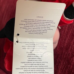 May include: Red plush toy with a black and white face. The toy is wearing a red and white striped shirt. The tag attached to the toy reads "The Walt Disney Company" and includes information about the company's address and contact information.