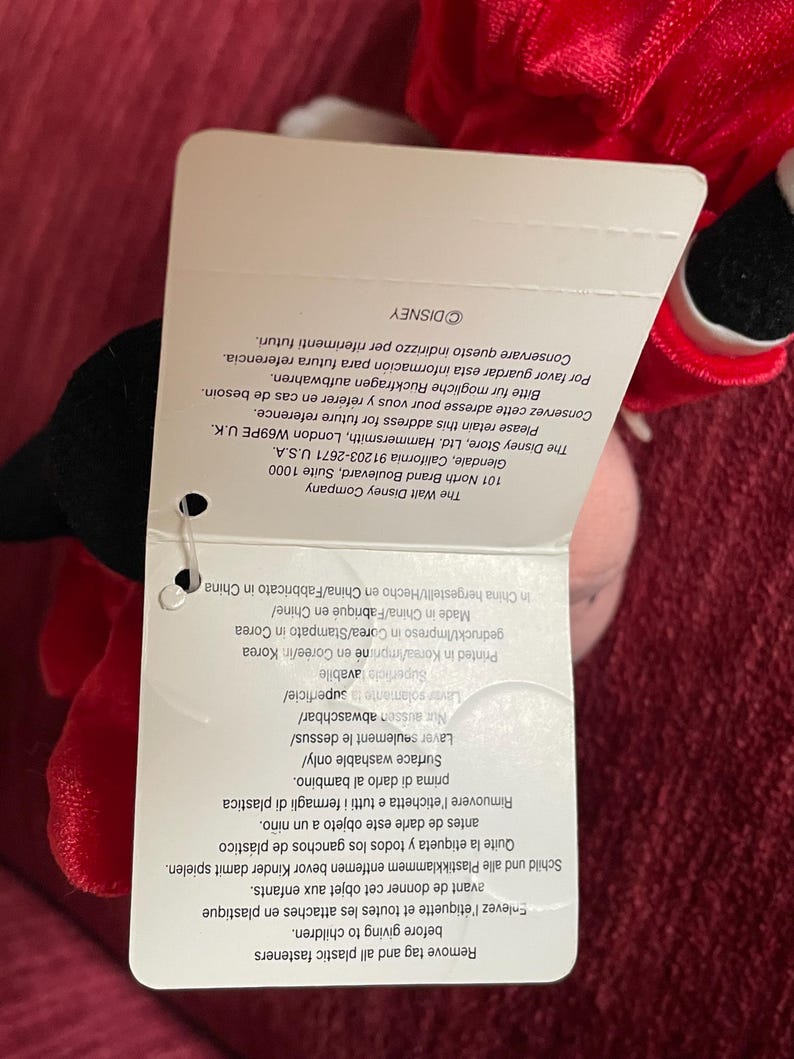 May include: Red plush toy with a black and white face. The toy is wearing a red and white striped shirt. The tag attached to the toy reads "The Walt Disney Company" and includes information about the company's address and contact information.