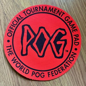 May include: A circular red game pad featuring the black text POG in the center. The outer edge includes the black text OFFICIAL TOURNAMENT GAME PAD and THE WORLD POG FEDERATION, with a small trademark symbol next to the central POG logo.