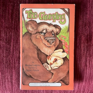 May include: A children's book titled "The Grumpling" with a brown bear hugging a sleeping rabbit. The cover has an orange border and text that reads "Good manners are an important part of daily life."