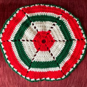 May include: A handmade, crocheted trivet in a circular shape. The trivet features concentric rings of red, white, and green yarn, with a scalloped edge. The central design is a red flower shape.