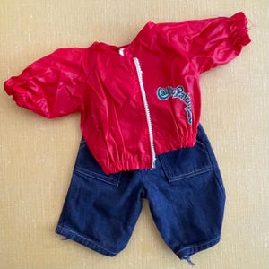 May include: A red nylon jacket with a white zipper and the text "Care Bears" in blue and green, paired with dark blue denim shorts. The jacket has short, puffy sleeves. The shorts have two front pockets.