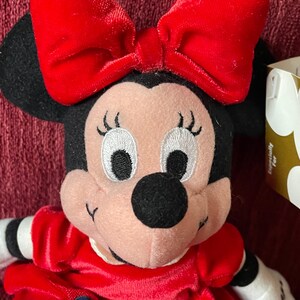 May include: Red and black plush toy of Minnie Mouse wearing a red velvet dress with a green holly and blue gem embellishment. She has a large red bow on her head and a white tag with the word "Minnie" on it.