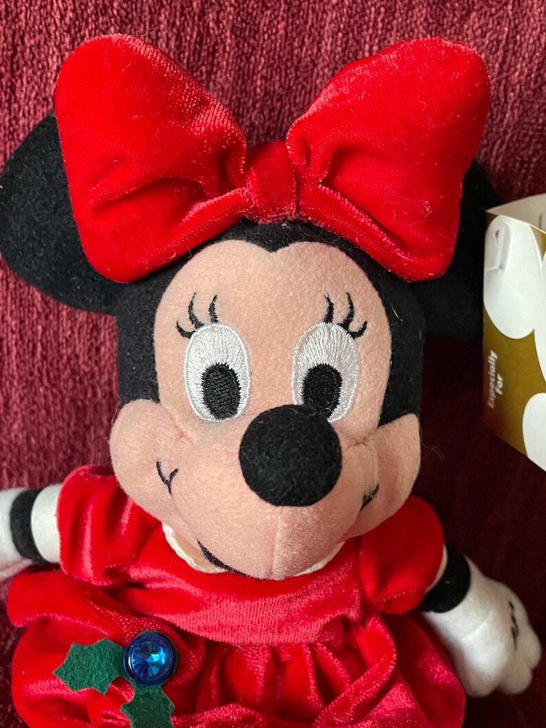 May include: Red and black plush toy of Minnie Mouse wearing a red velvet dress with a green holly and blue gem embellishment. She has a large red bow on her head and a white tag with the word "Minnie" on it.