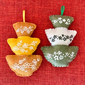 K&ouml;nnte beinhalten: Zwei Filz-Ornamente in Form von &uuml;bereinander gestapelten Schalen mit Blumendesigns. Das Ornament links ist gelb und braun, w&auml;hrend das Ornament rechts gr&uuml;n und wei&szlig; ist.