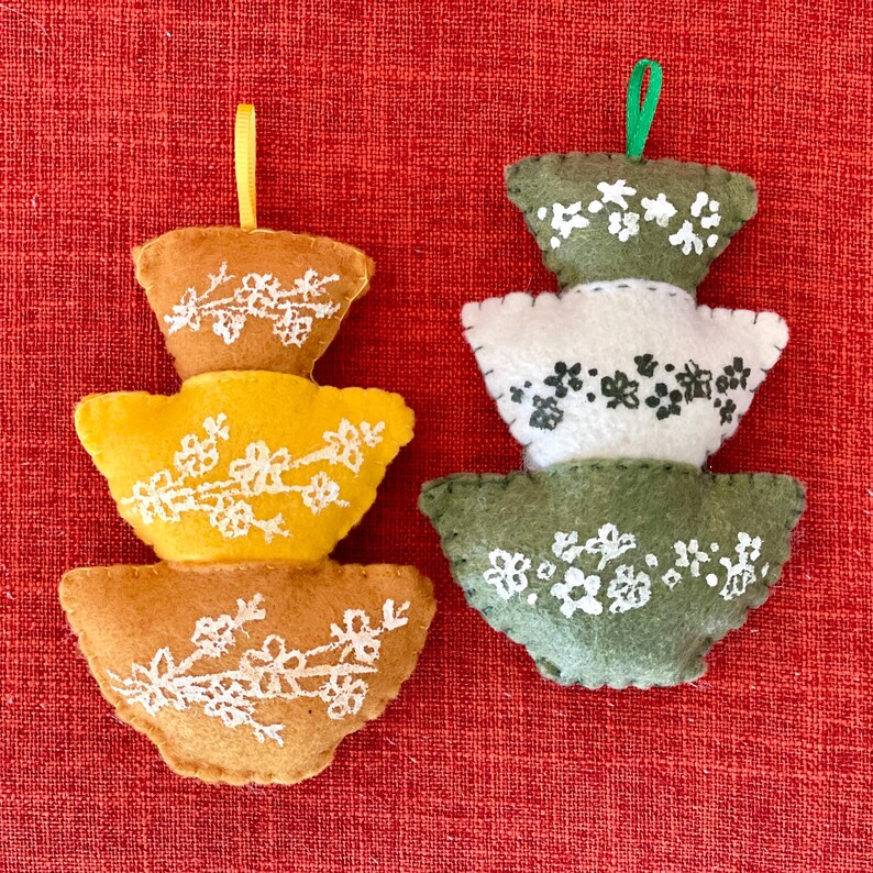K&ouml;nnte beinhalten: Zwei Filz-Ornamente in Form von &uuml;bereinander gestapelten Schalen mit Blumendesigns. Das Ornament links ist gelb und braun, w&auml;hrend das Ornament rechts gr&uuml;n und wei&szlig; ist.