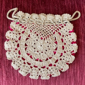 May include: A beige crocheted drawstring bag with a round, lacy design. The bag is on a red fabric background.