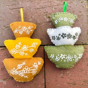 May include: Two felt ornaments shaped like stacks of bowls. The ornament on the left is yellow with white floral embroidery. The ornament on the right is green with white floral embroidery.