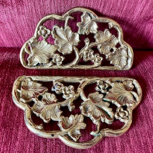 May include: Two antique gold-colored metal decorative pieces. Each piece has a unique shape with a floral and leaf design. The pieces are placed on a burgundy-colored fabric.