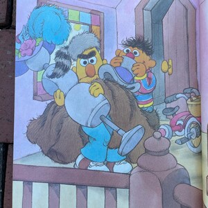 May include: Illustration from a children's book featuring Bert and Ernie. Bert, in blue pants and a brown coat, holds a large object. Ernie, in a striped shirt, assists. A bicycle is visible in the background.