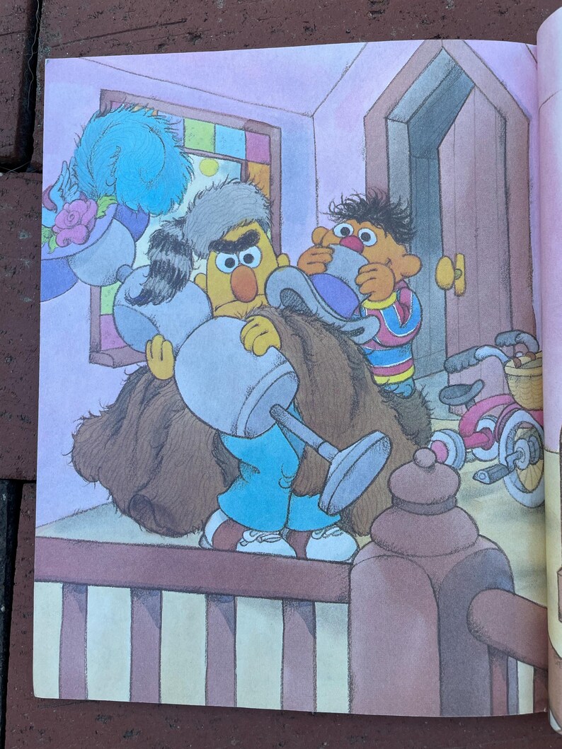 May include: Illustration from a children's book featuring Bert and Ernie. Bert, in blue pants and a brown coat, holds a large object. Ernie, in a striped shirt, assists. A bicycle is visible in the background.