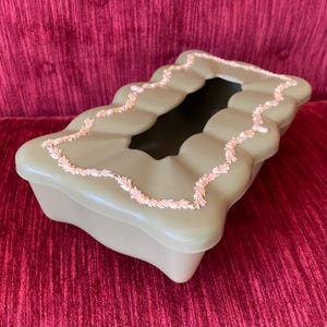 May include: A green tissue box cover with a decorative pink trim. The cover has a rectangular shape with rounded edges and a cutout for tissues.