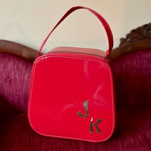 May include: A bright red, square-shaped vanity case with a glossy, patent-like finish and a single top handle. The front features two metallic, silver-toned letters, J and K, positioned in the lower right corner, with white stitching along the edges.