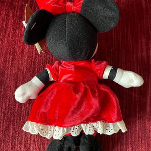 May include: A red velvet Minnie Mouse plush toy wearing a white lace trimmed dress and a red bow.