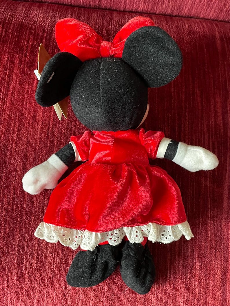 May include: A red velvet Minnie Mouse plush toy wearing a white lace trimmed dress and a red bow.