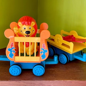 May include: A toy train set with a lion in a cage car and a flatbed car. The lion is orange and yellow with a red mane. The train cars are blue, orange, and yellow.