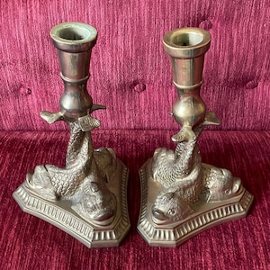 May include: A pair of silver-toned candle holders. Each holder features a dolphin design, with the dolphin's body forming the base and supporting the candle cup. The holders have a detailed, antique appearance, and are set against a burgundy background.