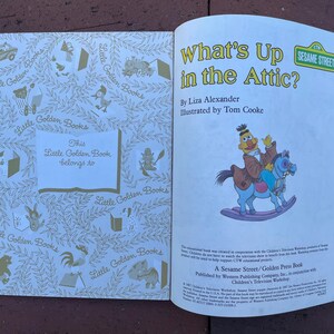 May include: An open Little Golden Book titled "What's Up in the Attic?" featuring a Sesame Street character riding a rocking horse. The book's cover is white with gold illustrations and the text "This Little Golden Book belongs to".
