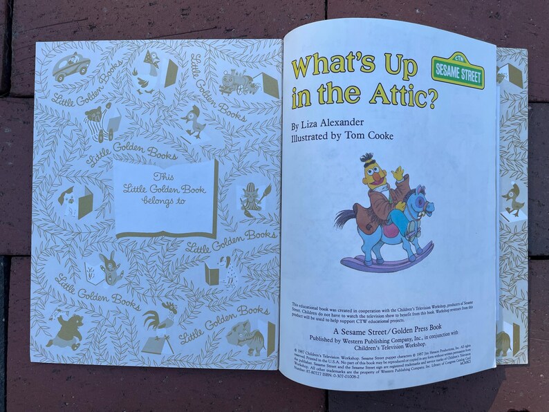 May include: An open Little Golden Book titled "What's Up in the Attic?" featuring a Sesame Street character riding a rocking horse. The book's cover is white with gold illustrations and the text "This Little Golden Book belongs to".