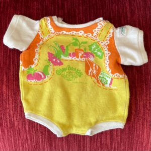 May include: A vintage Cabbage Patch Kids doll outfit. The one-piece garment is yellow with orange trim and white sleeves. The front features a colorful garden scene with the Cabbage Patch Kids logo.