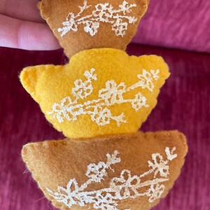 K&ouml;nnte beinhalten: Ein Stapel aus drei Filz-Ornamenten in Form von Vasen. Die Vasen sind braun und gelb mit wei&szlig;en gestickten Blumenmustern.