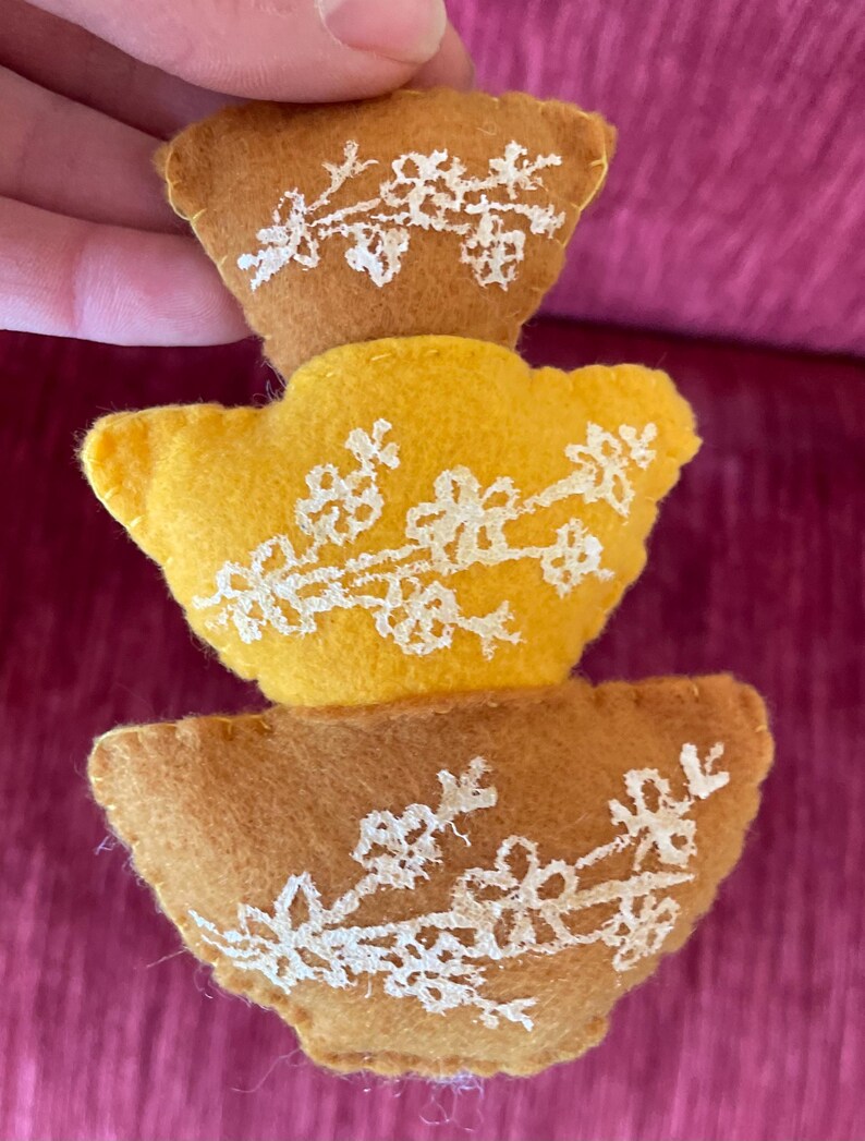 K&ouml;nnte beinhalten: Ein Stapel aus drei Filz-Ornamenten in Form von Vasen. Die Vasen sind braun und gelb mit wei&szlig;en gestickten Blumenmustern.