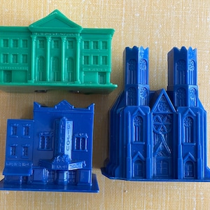 May include: Three plastic building miniatures: a green classical building, a blue theater with "BIJOU THEATRE" text, and a blue cathedral. The buildings are on a yellow background.